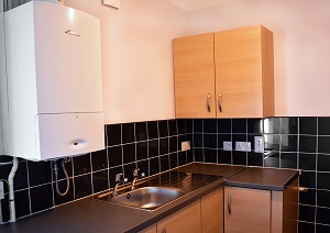 A kitchen in a Council property with a boiler, sink, cabinets and black tiles.