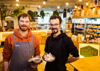 Essen store owners with 2 small plates of food in their shop